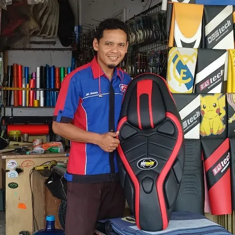 Pria berseragam biru-merah memegang jok motor hitam bergaris merah di toko yang penuh sarung jok motor